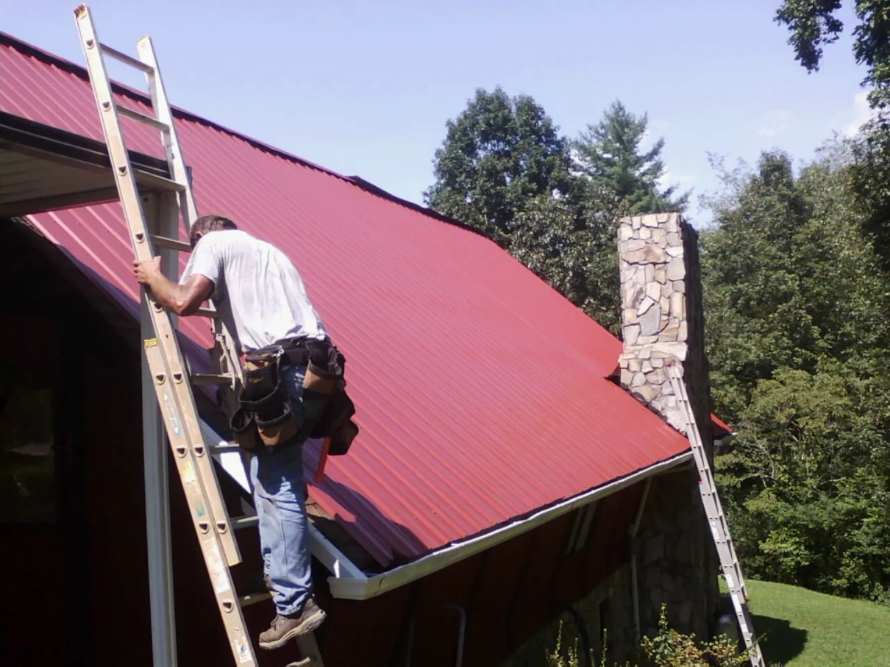 Roofer on a ladder beside a red metal roof and stone chimney — Soffit & Fascia Repair by Roofing Brothers in Strongsville
